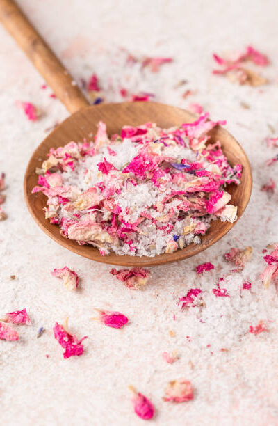 See salt with with dried flower petals in wooden scoop on pink marble table
